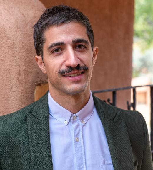 Headshot of smiling man with mustache, green blazer and light blue shirt against adobe wall.